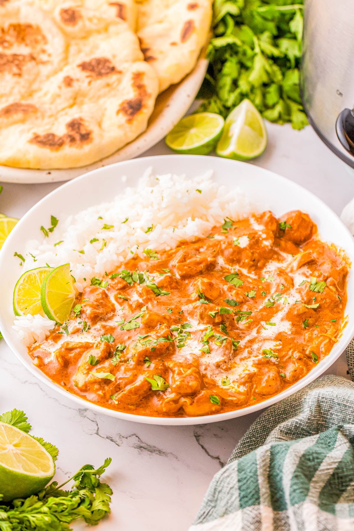 A bowl of chicken tikka masala with white rice, garnished with cilantro and lime wedges, served with naan bread on the side.