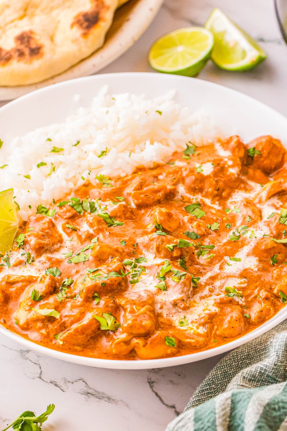 A bowl of chicken tikka masala garnished with cilantro, served with white rice, lime wedges, and naan bread.