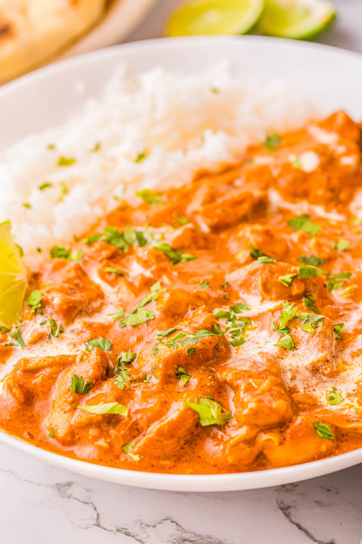 A bowl of chicken tikka masala garnished with cilantro, served with a side of white rice and lime wedges.