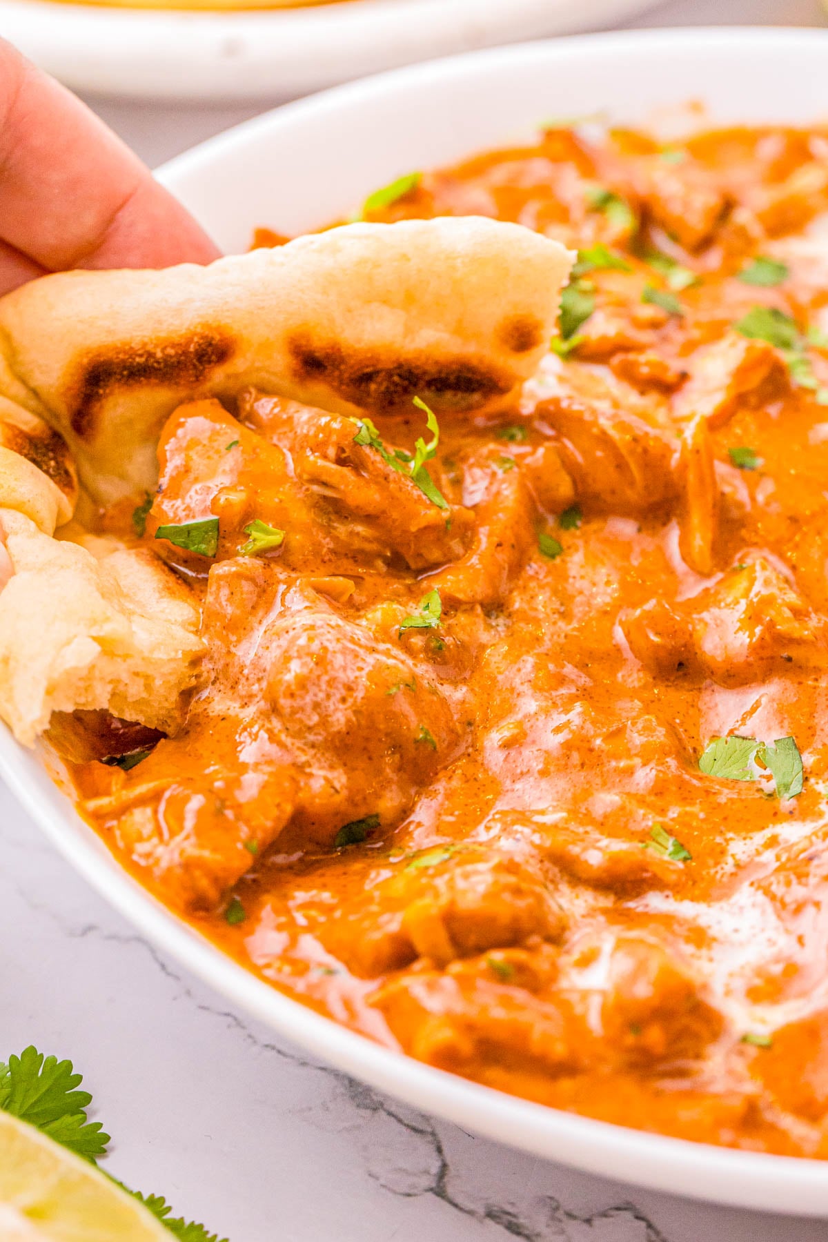A piece of naan dipping into a bowl of creamy, orange-colored curry topped with fresh herbs.
