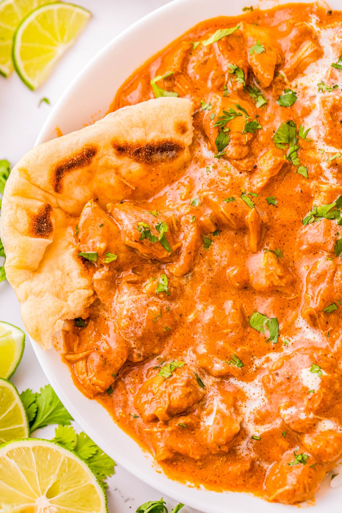 A bowl of creamy chicken curry garnished with cilantro, served with a piece of naan bread and lime wedges.