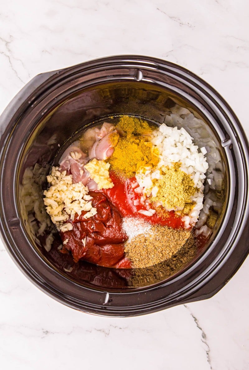 Ingredients for a meal, including raw chicken, diced onion, minced garlic, tomato paste, and various spices, arranged in a slow cooker.