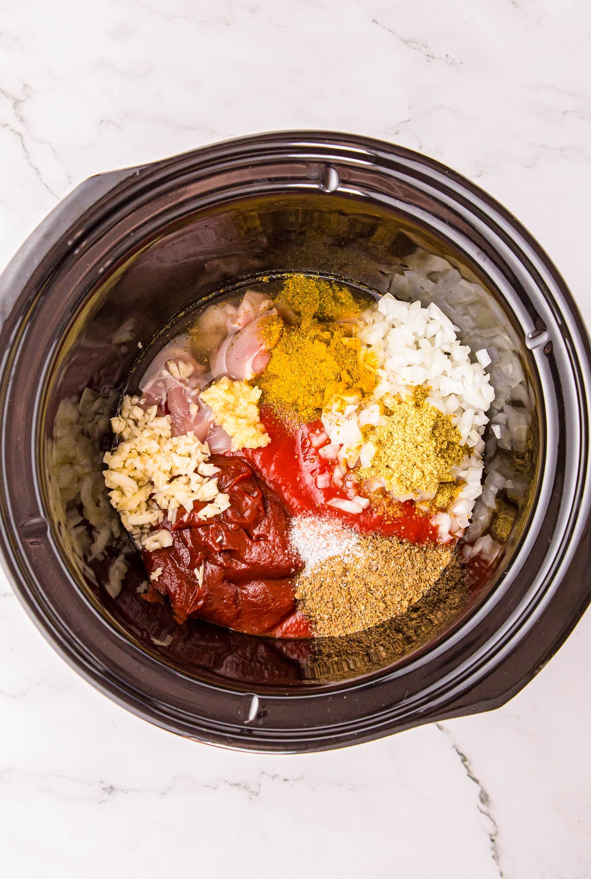 Ingredients for a meal, including raw chicken, diced onion, minced garlic, tomato paste, and various spices, arranged in a slow cooker.