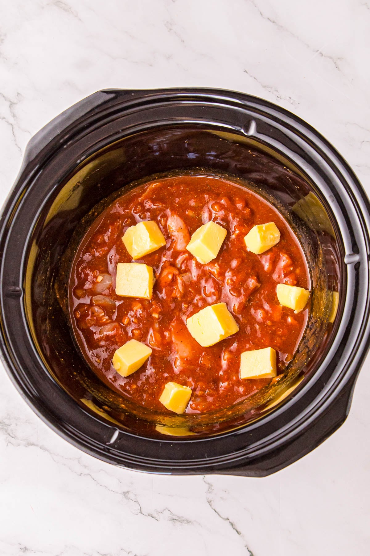 Slow cooker filled with a red sauce topped with several cubes of butter.