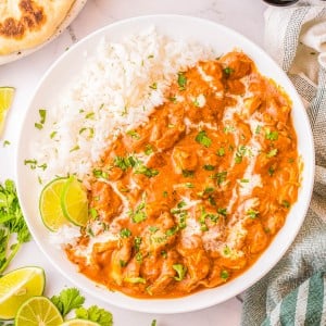 A bowl of chicken tikka masala served with white rice, garnished with cilantro and a drizzle of cream, with lime wedges and naan bread on the side.