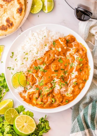 A bowl of chicken tikka masala served with white rice, garnished with cilantro and a drizzle of cream, with lime wedges and naan bread on the side.