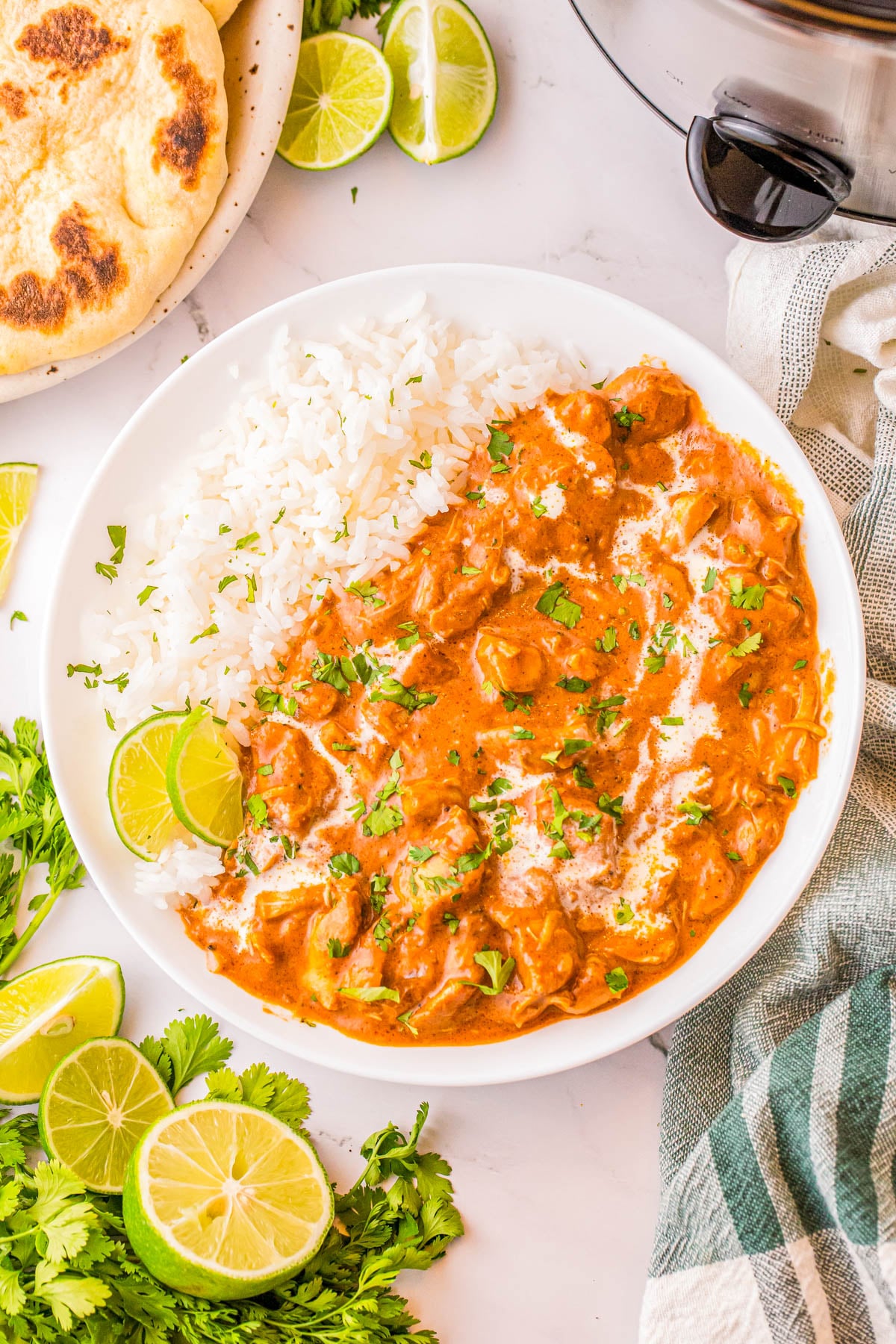 A bowl of chicken tikka masala served with white rice, garnished with cilantro and a drizzle of cream, with lime wedges and naan bread on the side.