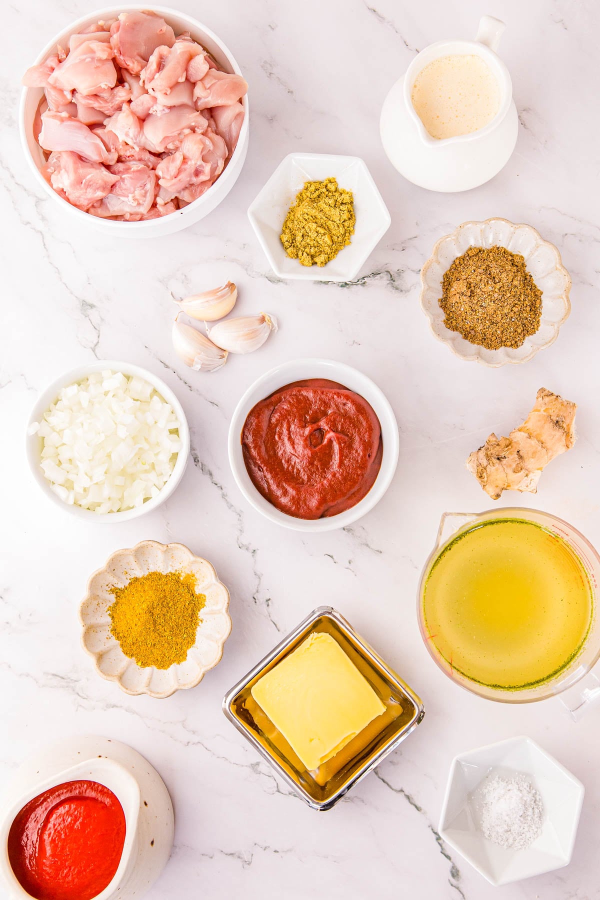 Ingredients for a recipe arranged on a white marble surface: raw chicken, onions, garlic, ginger, tomato paste, butter, cream, broth, and various spices.